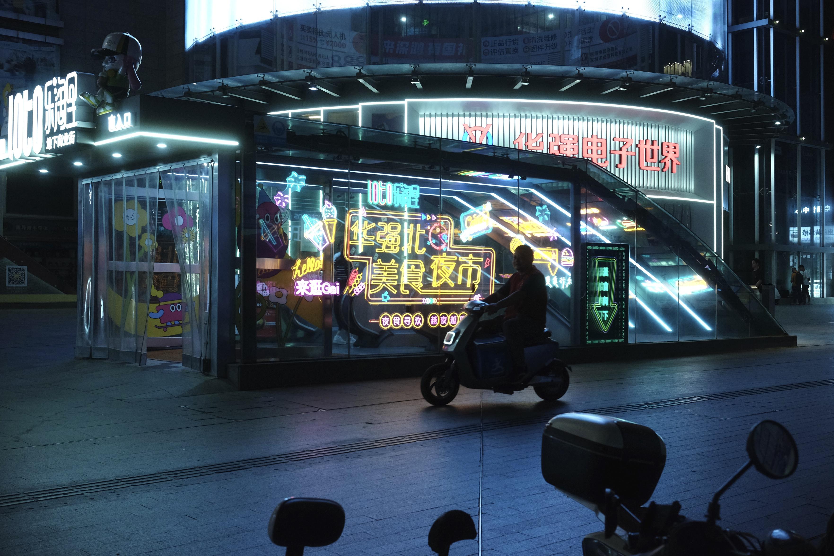 A nighttime photo of an entrance to some form of train station or something similar. It is laced with neon signs of Chinese characters and food choices. Infront a person is scooting by on an almost futuristic vehicle. A round building with red Chinese characters frames the background A nighttime photo of an entrance to some form of train station or something similar. It is laced with neon signs of Chinese characters and food choices. Infront a person is scooting by on an almost futuristic vehicle. A round building with red Chinese characters frames the background