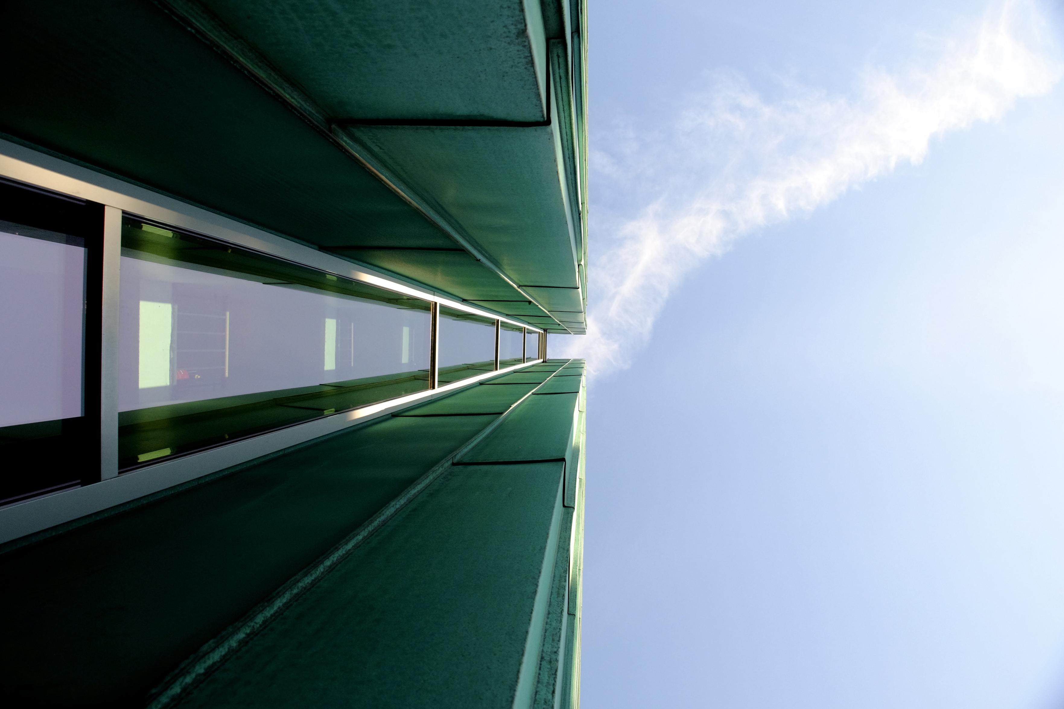 A photo up two green pillars that are connected by a long strip of windows. The pillars cut the image right in the middle, leaving the right half empty except for some lightly clouded sky A photo up two green pillars that are connected by a long strip of windows. The pillars cut the image right in the middle, leaving the right half empty except for some lightly clouded sky