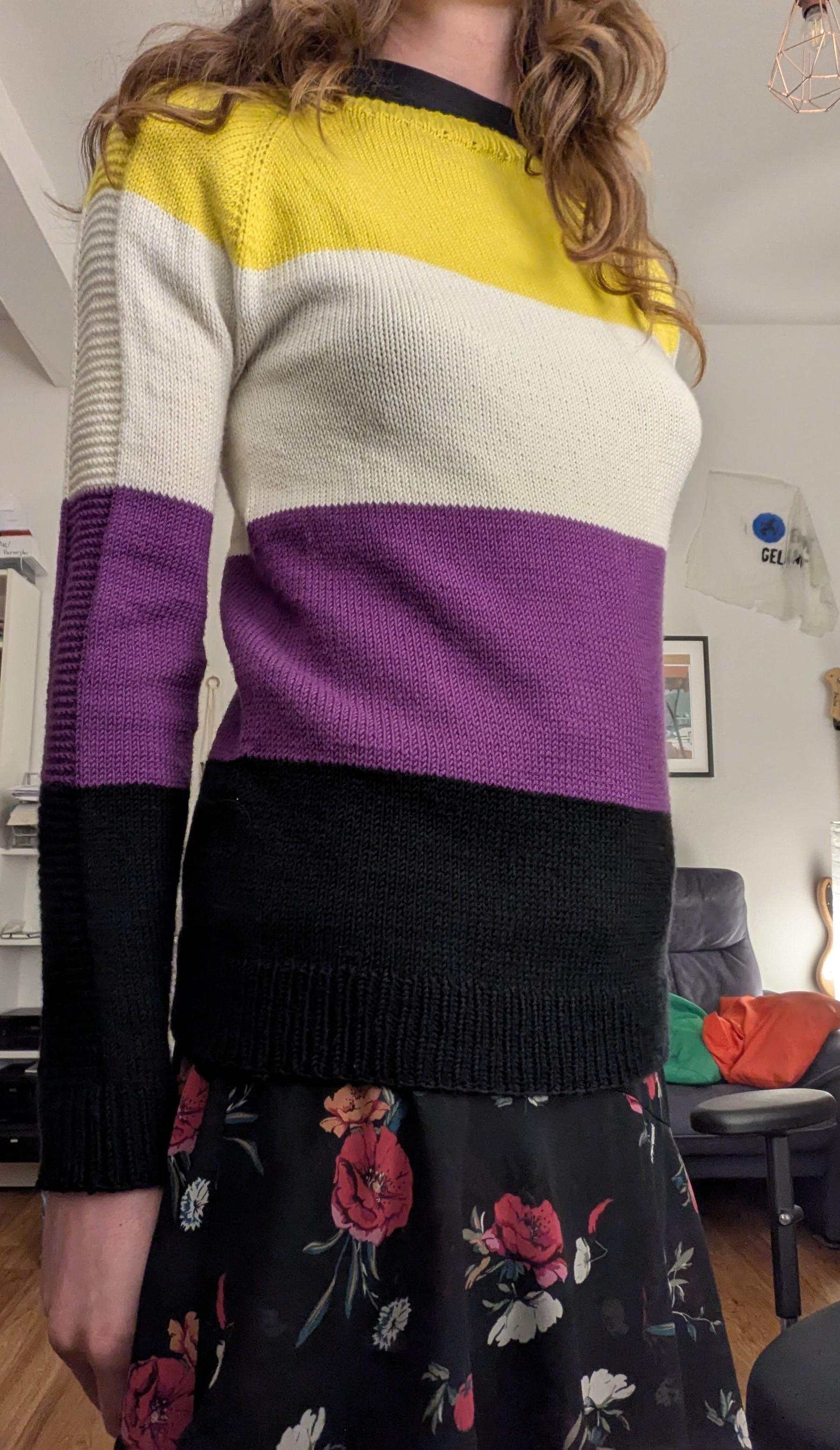 person wearing a sweater in the four stripes of the nonbinary flag (yellow, white, purple, black)
brown wavy hair drapes down onto the shoulders.
There's a raglan pattern on the outside of the arm