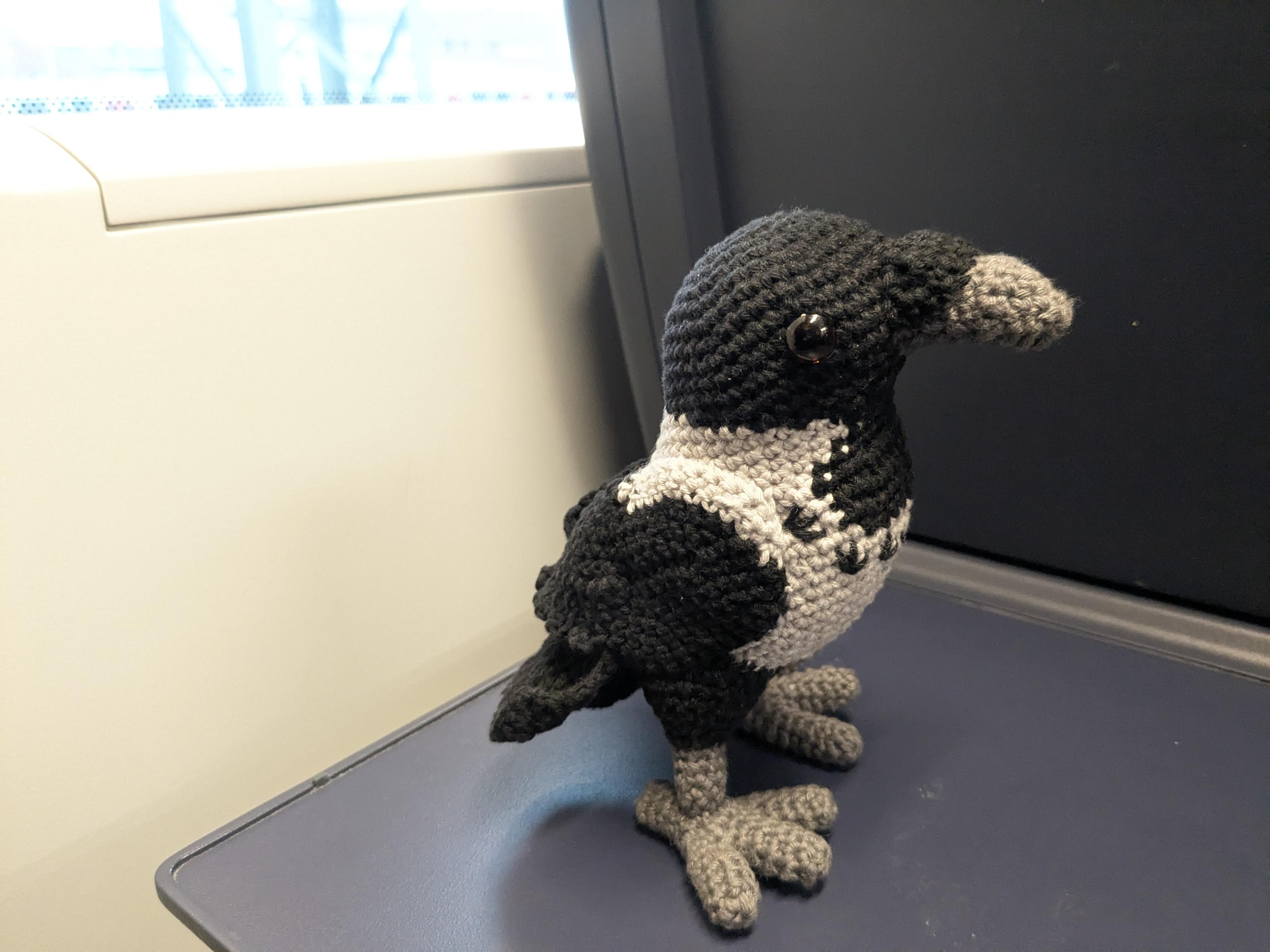 A crochet hooded crow standing up. It has a black head with brown button eyes, a dark grey beak, an off-white torso and wings and legs and tail feathers that are black again. It's standing on stuffed rounded legs and feet on a table on a train.