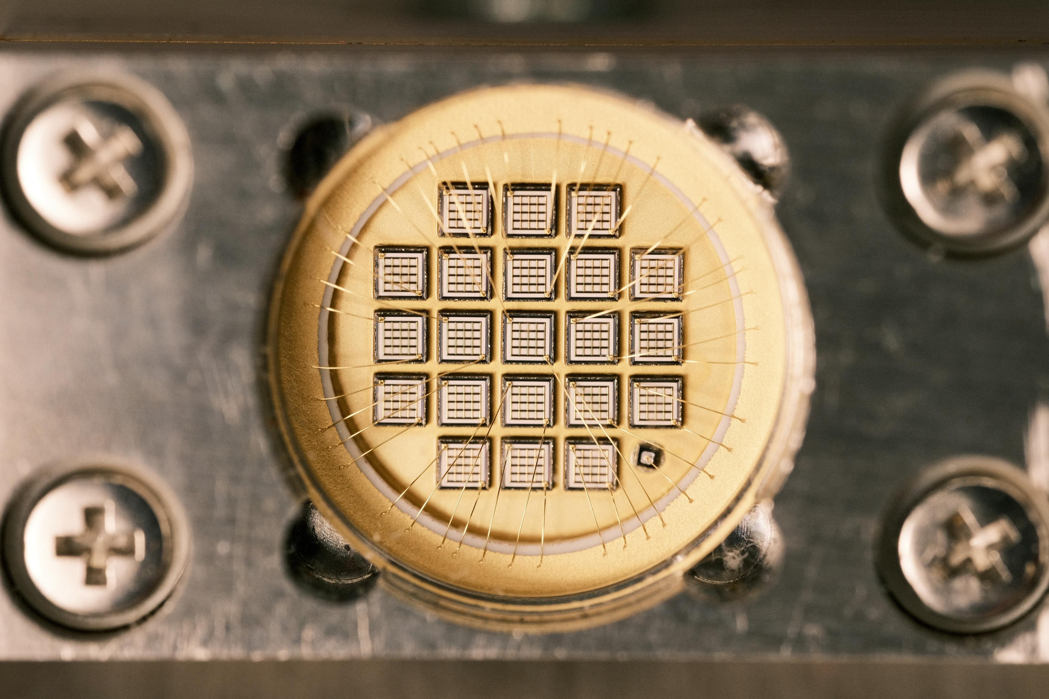 A closeup of the array of dies. It’s 21 small dies that each have a small array of four by four squares in them. A bunch of golden bond-wires escape to the side where they connect to a golden ring. The golden ring is isolated from the golden backplate by another white ring