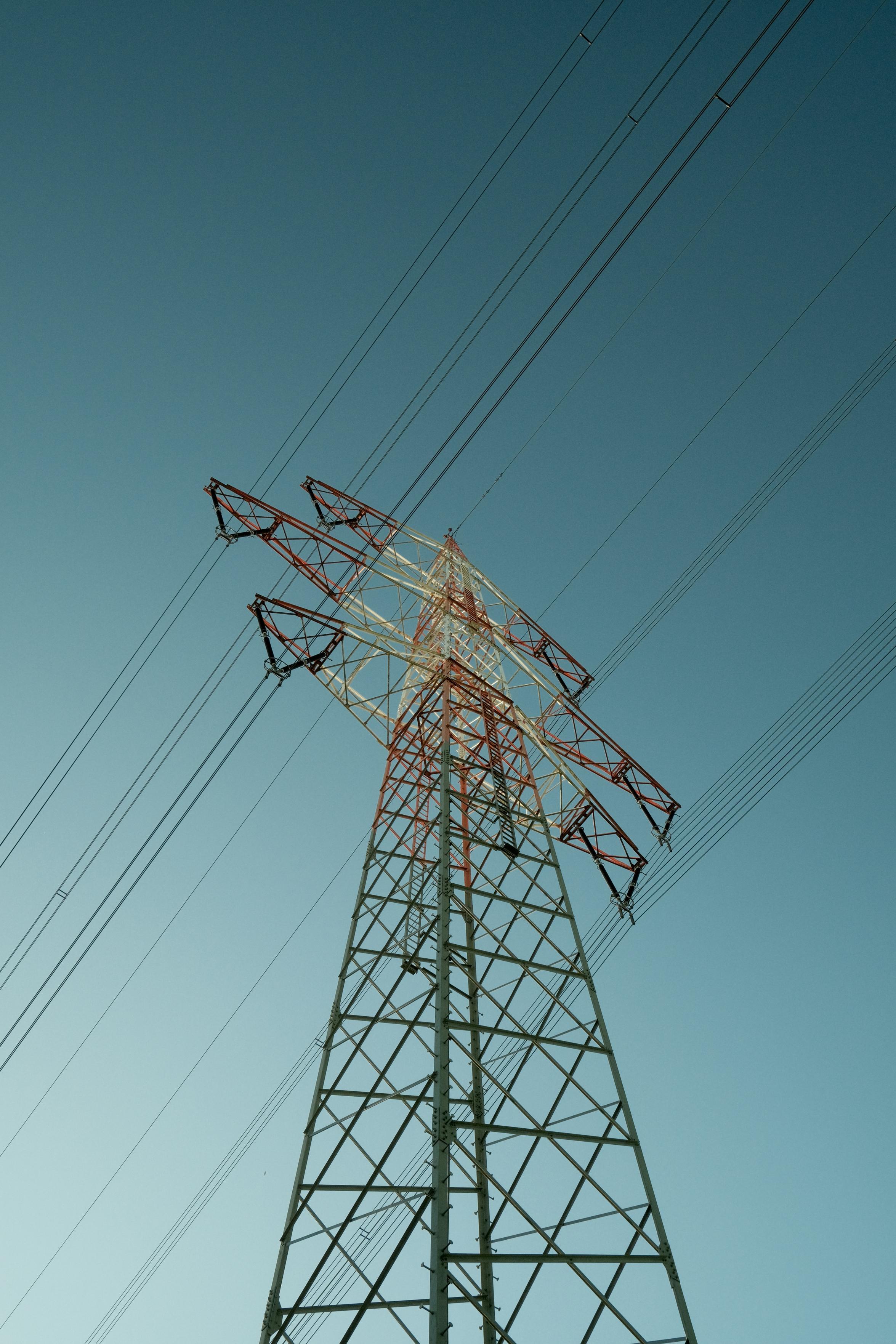 A photo up a tall pylon. It’s half grey and half red-white coloured. The top is illuminated, probably sunlight. Powerlines run through the image, cutting the dark aquamarine sky