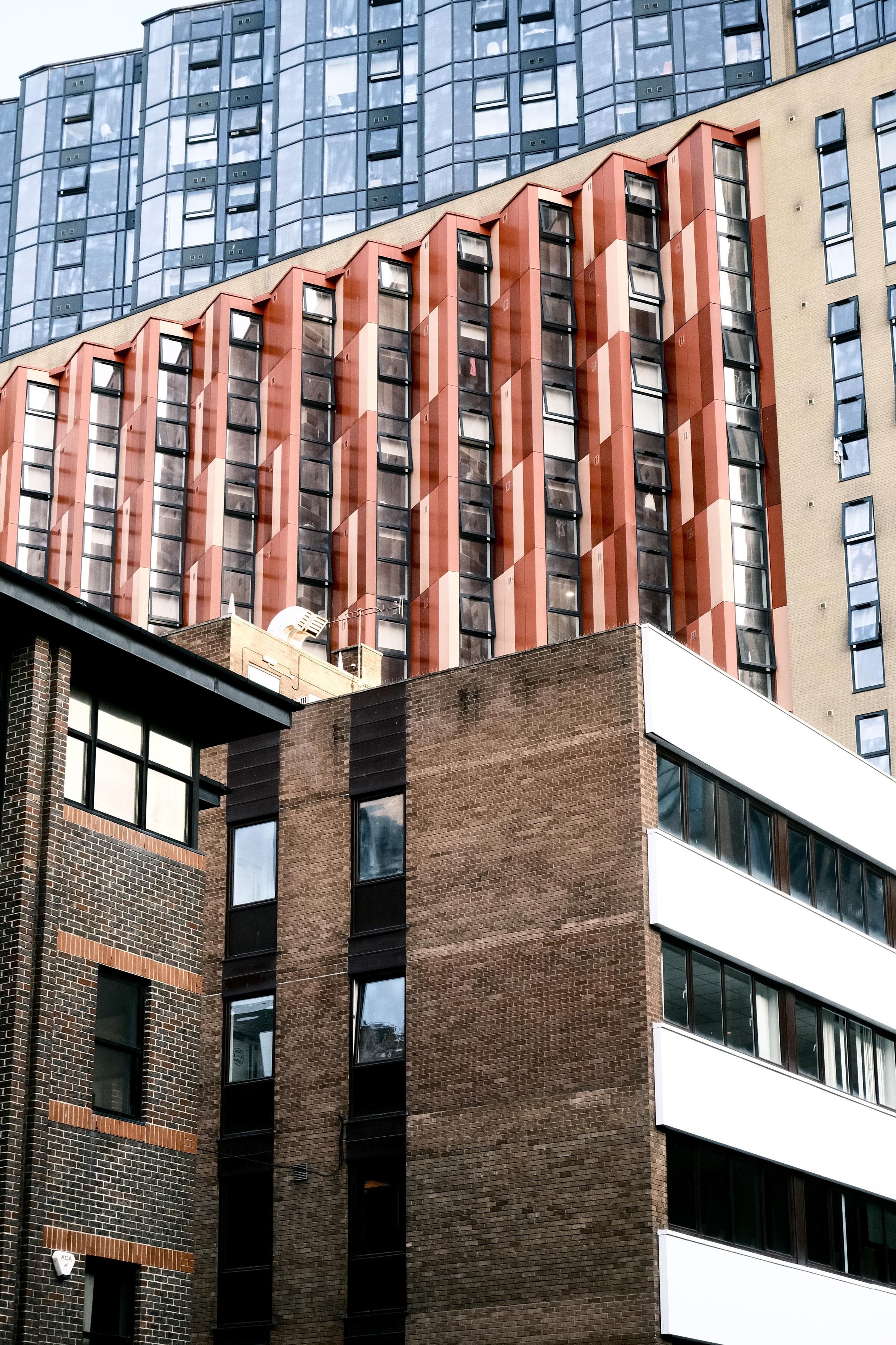 A layered image of several buildings. The closest one is made of brick, more traditional, an office maybe. The one behind is a more modern block living situation made of red ripped outset windows. They all face away from the building at maybe sixty degrees. Above a blue block of glass fills where the sky should be. More apartments maybe, or offices