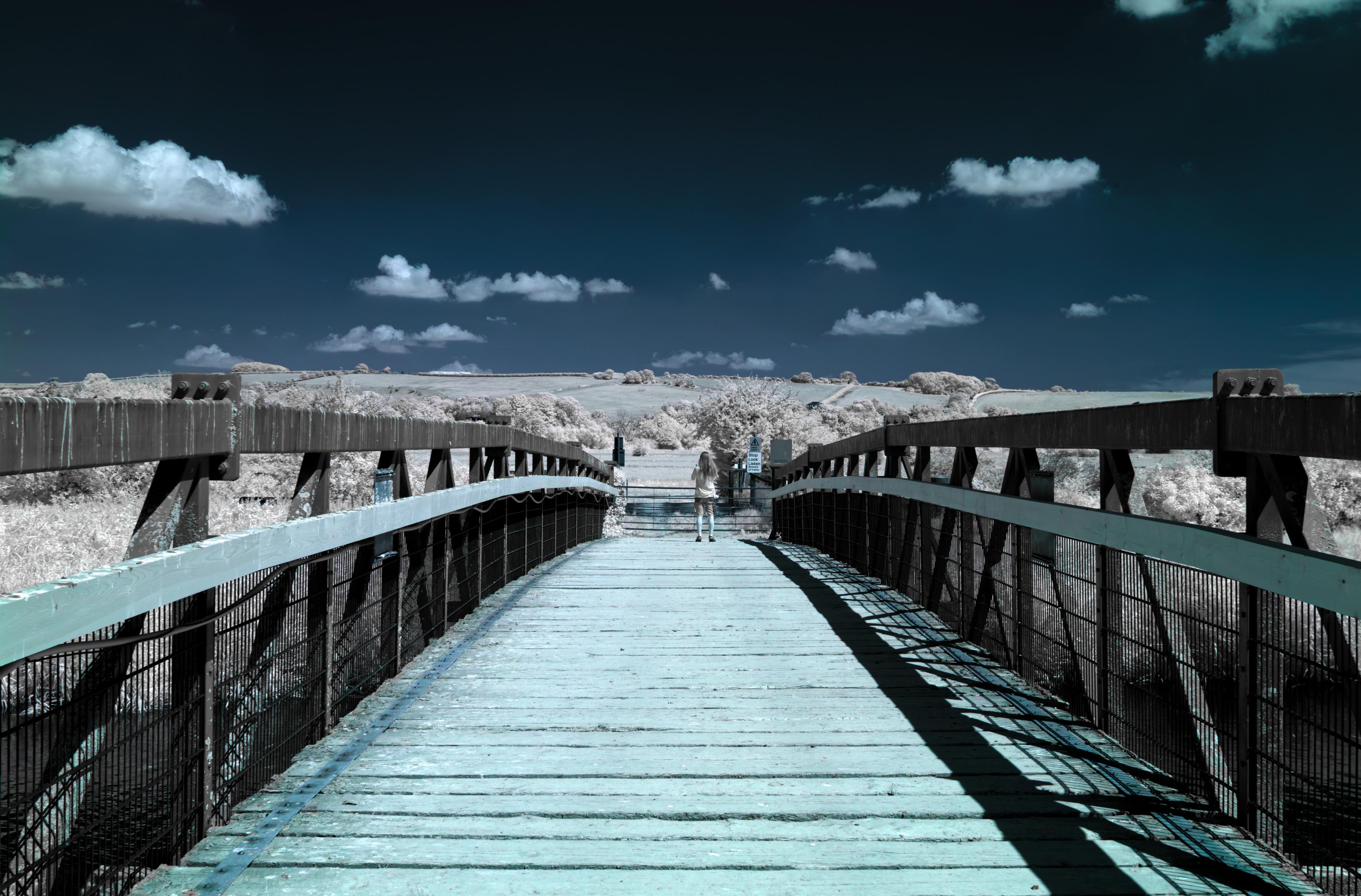 A photo over a wooden bridge. It is long, somewhat wide and not decorated. A gentle hilly landscape emerges on the other side. The sky is dark blue with light contrasty clouds. The bridge is ash grey, the landscape creamy white. A female is standing on the end of the bridge, looking into the landscape. It’s an infrared photo